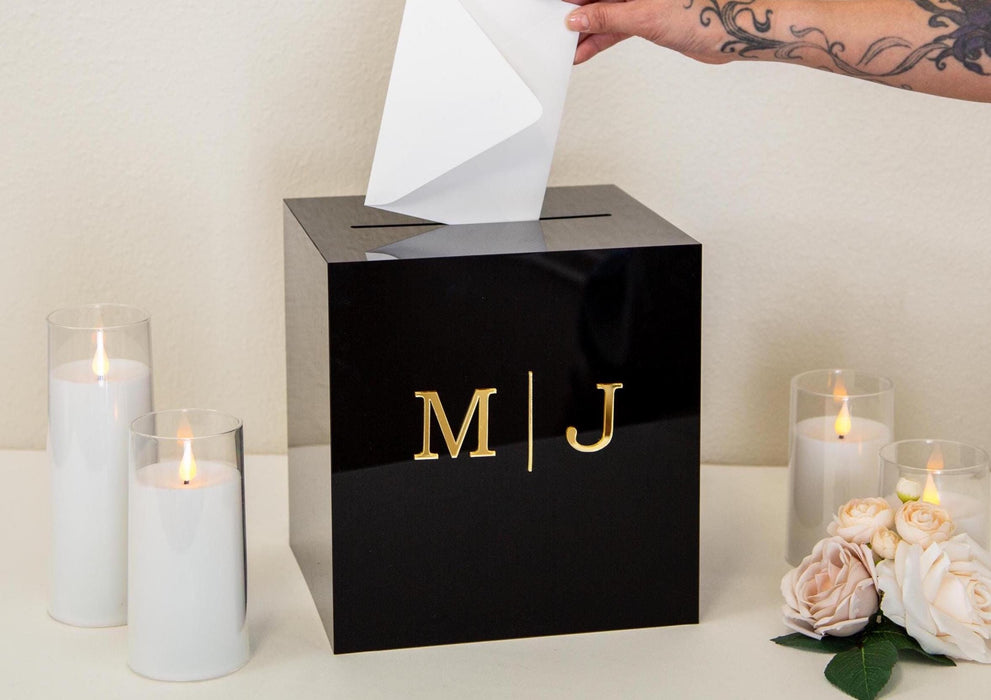 May include: A black acrylic wedding card box with a gold inscription that reads 'M|J'.