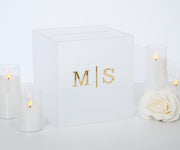 May include: A white acrylic box with a gold inscription that reads 'M|S'. The box is surrounded by four white candles and a white rose.