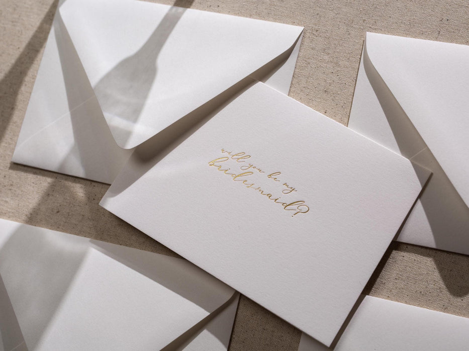May include: A white card with gold foil lettering that says 'Will you be my bridesmaid?'
