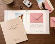 May include: A beige photo album with gold accents, open to display photos and a pink envelope. The cover reads "Reasons Why I love you" with the names "Richard & Mimi" below. A vintage camera and ring are also visible.