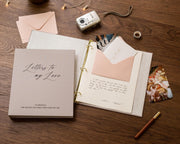 May include: A beige scrapbook titled "Letters to my Love" with gold binder rings, open to reveal a handwritten letter and a pink envelope. A small camera, photos, and a pen are also on the wooden surface.