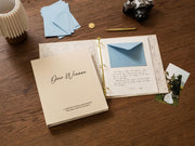 May include: An open scrapbook with the words "Dear Winona" on the cover, along with the text "A collection of letters and memories from those who love you most." Inside, a blue envelope and handwritten letter are visible. A vintage camera and blue envelopes are also present.