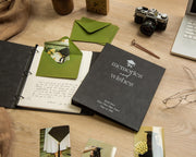 May include: A black binder with the words "memories and wishes" in white, a graduation cap icon, and the name "Sarah Jones" and "Class of 2026". The binder is open, revealing handwritten text and a green envelope. A vintage camera, pen, and photos are also present.