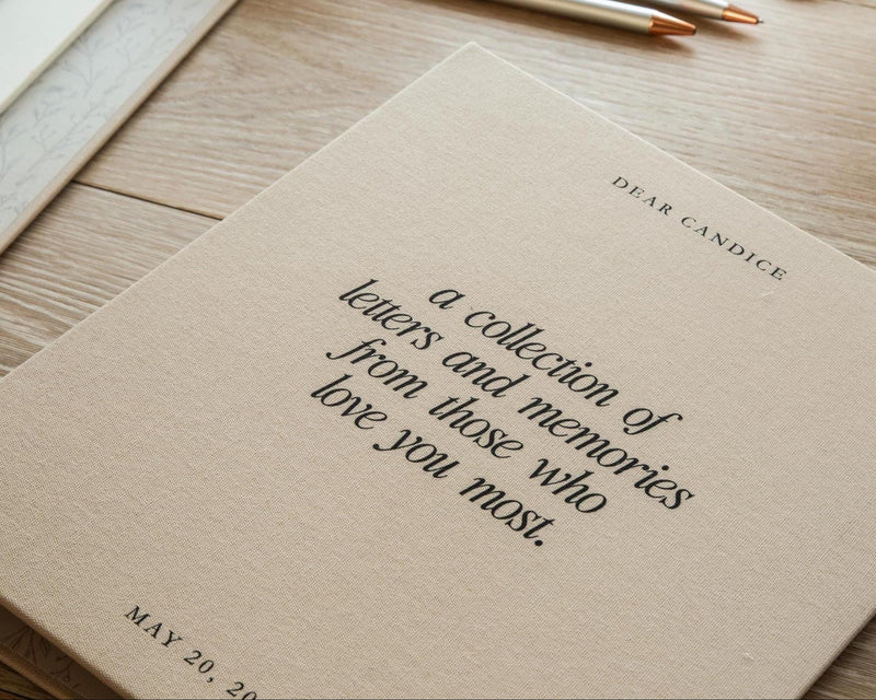 May include: A beige fabric-covered book with black text that reads DEAR CANDICE, a collection of letters and memories from those who love you most, and MAY 20, 20. The cover has a textured surface and is positioned at an angle on a wooden surface.