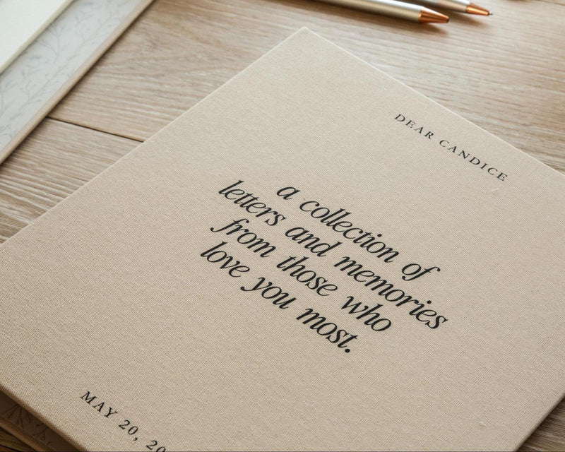 May include: A beige fabric-covered book with black text that reads DEAR CANDICE, a collection of letters and memories from those who love you most, and MAY 20, 20. The cover has a textured surface and is positioned at an angle on a wooden surface.