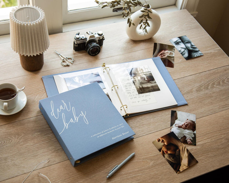 May include: A dusty blue three-ring binder photo album with the text "dear baby" and "A collection of letters & photos to open at different moments in your life" printed on the cover, alongside loose photographs, a camera, and a pen.