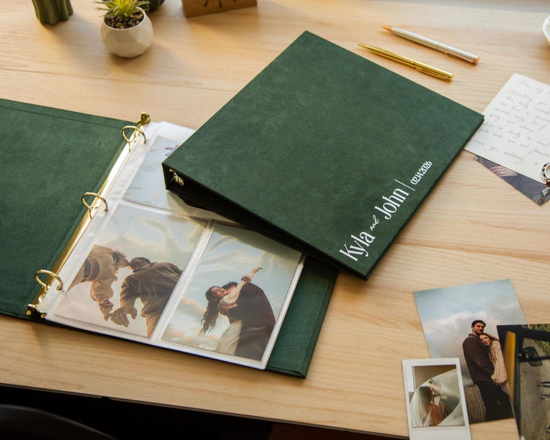 May include: A dark green photo album with gold-colored rings, open to display wedding photos. The cover is embossed with the names "Kyla and John" and the date "02.11.2026" in white. The album is on a wooden surface.